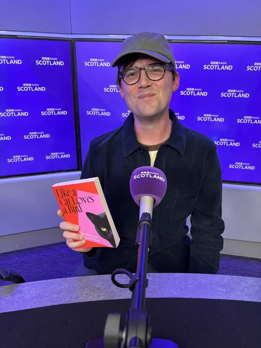 Me, behind a microphone in the BBC Radio Scotland studio. I’m holding a copy of my book, Like a Cat Loves a Bird: The Nine Lives of Muriel Spark
