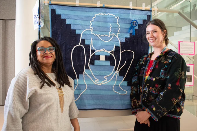 Two woman standing next to a quilt constructed from blue medical textiles recovered from the Parnassus Operating room including scrub caps, bunny suits, and disposable scrub jackets. An image of a birthing mother is overlaid made out of braided strips of material, evoking Black hair braiding techniques.