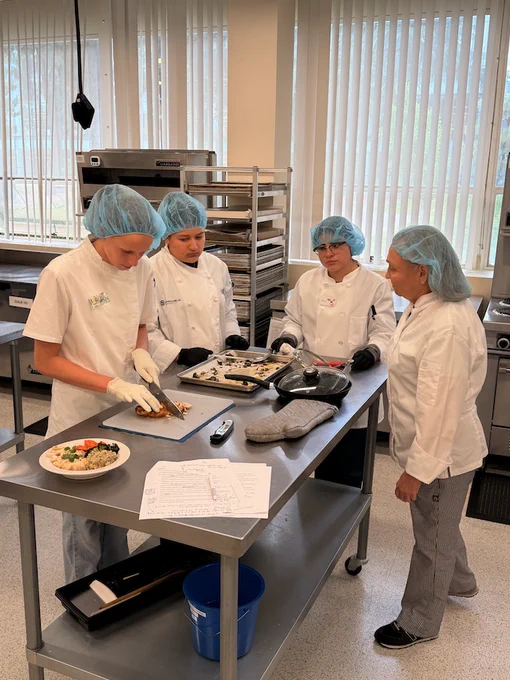 Four people in a commercial kitchen are preparing food. They are wearing white chef coats and blue hair nets. One person is slicing food on a cutting board while others observe. A bowl of ingredients and a clipboard with paper are on the stainless steel table.