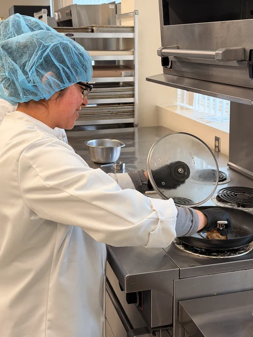 A person in a white lab coat and blue hair net is cooking on a stovetop in a professional kitchen. They are lifting the lid of a frying pan with steam visible. Stainless steel bowls and equipment are in the background.