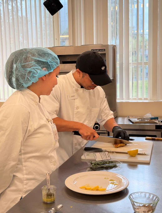Two people in a kitchen, wearing white chef uniforms. One is slicing food on a cutting board while the other watches. A plate with garnish and lemon slices is on the counter. A large oven and windows are in the background.