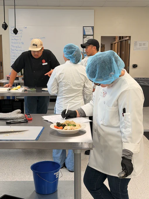 A group of individuals in a kitchen setting, wearing protective hair covers and lab coats, engage in a discussion. A plate of food is on the table. The room has a whiteboard and kitchen equipment, suggesting a culinary class or workshop.