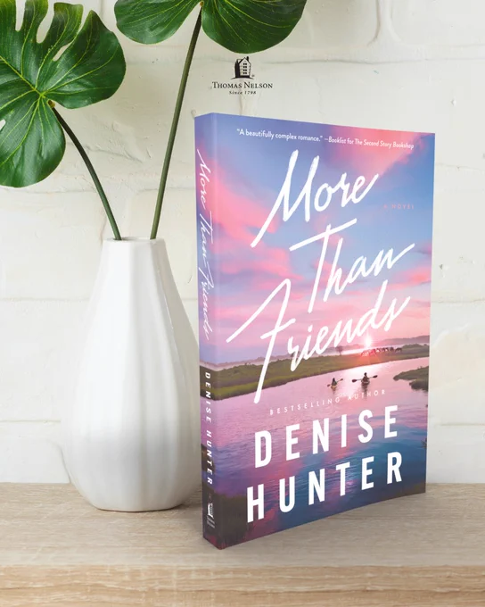 The image shows a book titled "More Than Friends" by Denise Hunter. It is resting upright on a wooden surface next to a white vase holding a large green plant. The background is a light brick wall. The cover features a serene landscape with pink and blue skies over water.
