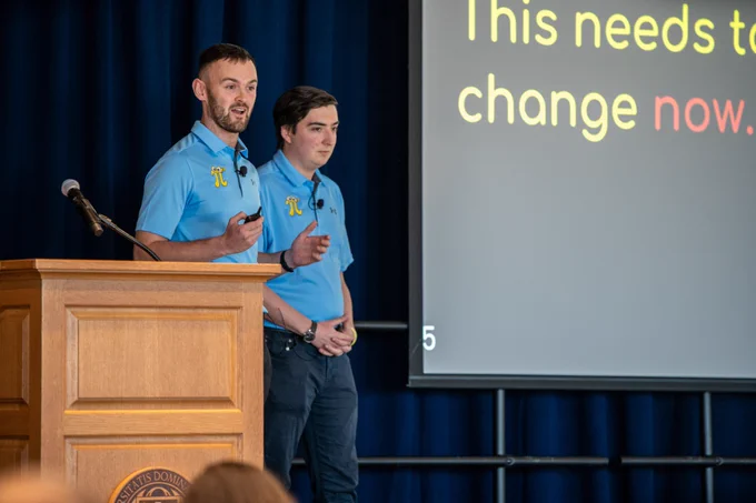 Two men in light blue polo shirts present at a podium.
