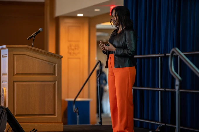 A woman wearing orange pants and a leather jacket talks and gestures with her hands.