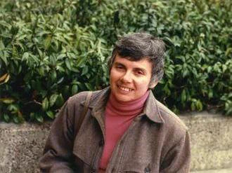 Photograph of mathematician Louise Hay wearing a brown collared jacket and a red turtleneck sweater sitting outdoors in front of dense green foliage, with a concrete ledge behind her.