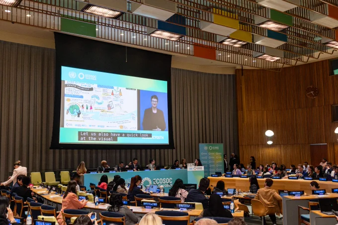 Conference room with attendees watching a large screen showing a speaker and an illustrated presentation at an official event.