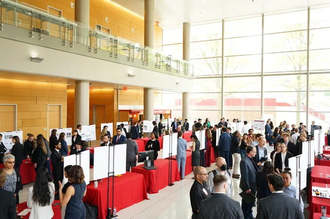 A busy indoor poster session event in a large hall with tall windows, featuring numerous people interacting around tables with red tablecloths. Display boards are set up on the tables, and the atmosphere is lively and professional.