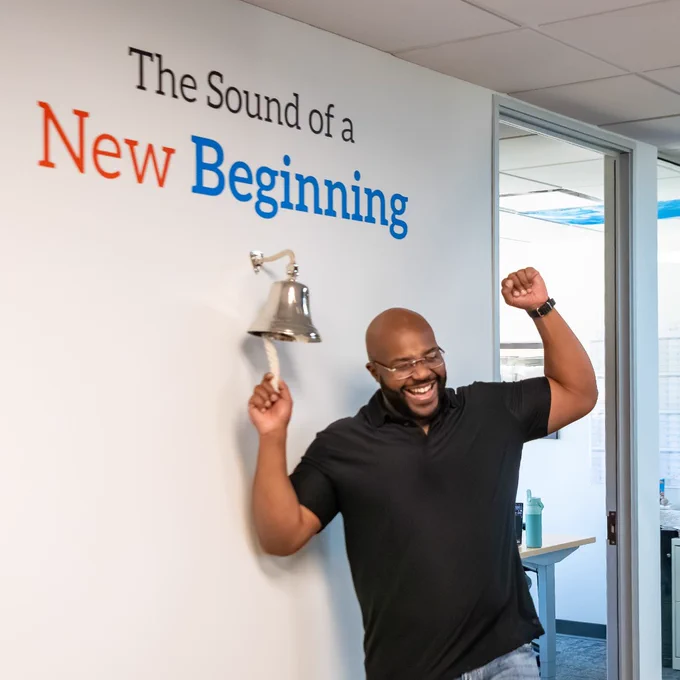 Man cheering and ringing a silver bell with the phrase 'The Sound of a New Beginning' on the wall above it in an office setting.