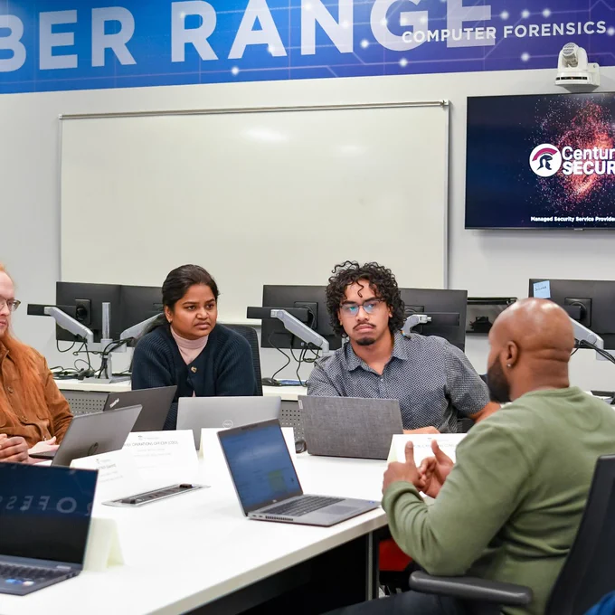 Group of diverse professionals collaborating around a table with laptops in a Cyber Range computer forensics lab.