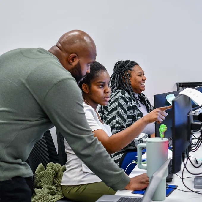 A man in a green sweater leans over to assist two women working at computers in a modern office setting.