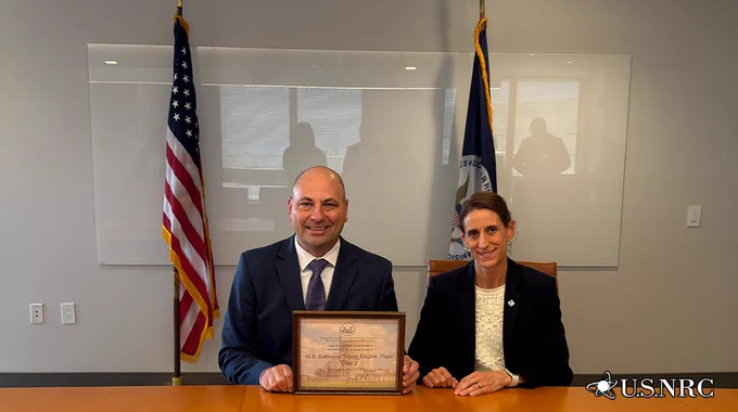 Two officials seated at a table holding a framed award with U.S. and NRC flags behind them in a formal office setting.