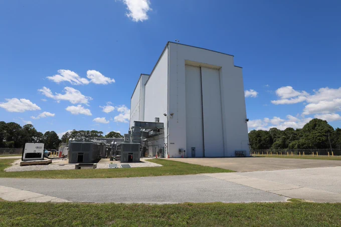 Image shows sunny, blue skies with a few wispy Cirrus clouds outside the NASA Kennedy’s Payload Hazardous Servicing Facility (PHSF). The building is a grayish color with a nearby driveway and a small door for people to enter and huge doors for spacecrafts and instruments to enter. Photo credit: NASA/Kim Shiflett