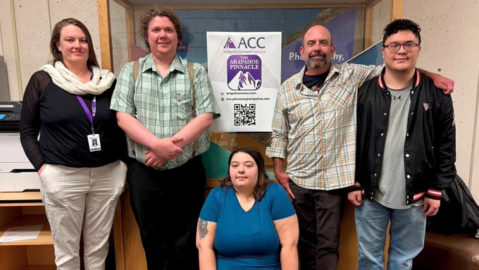Five staff members and faculty sponsors from the Arapahoe Pinnacle are pictured posing in front of a display case on  the fourth floor of the Arapahoe Community College Littleton Campus.