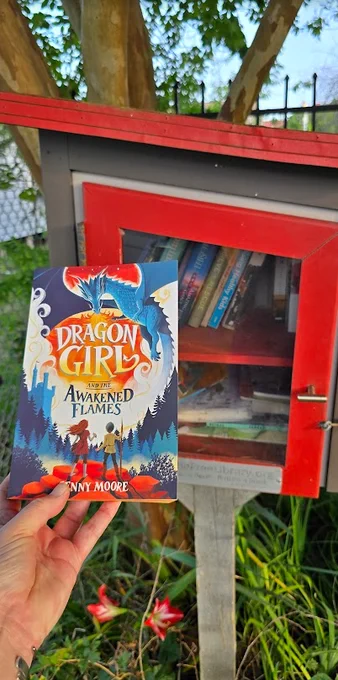 Book being held up in front of a Little Free Library. 