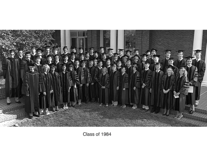 A formal group portrait of the Heritage College Class of 1984 wearing graduation caps and gowns in front of a brick building.