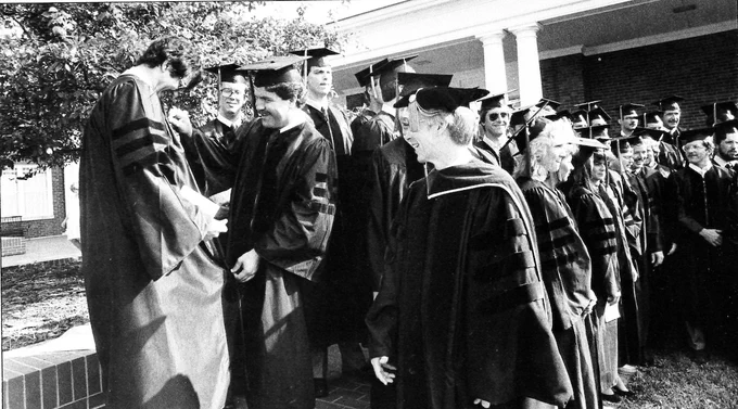 A black-and-white photo of graduates in academic regalia smiling and talking outside at their graduation ceremony.