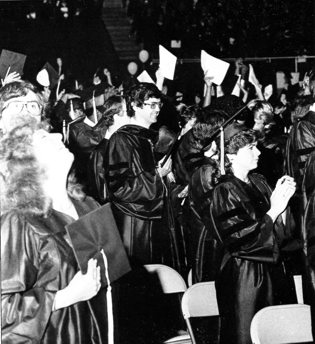 A candid black-and-white photo of graduates celebrating and raising their programs in the air during a commencement ceremony.