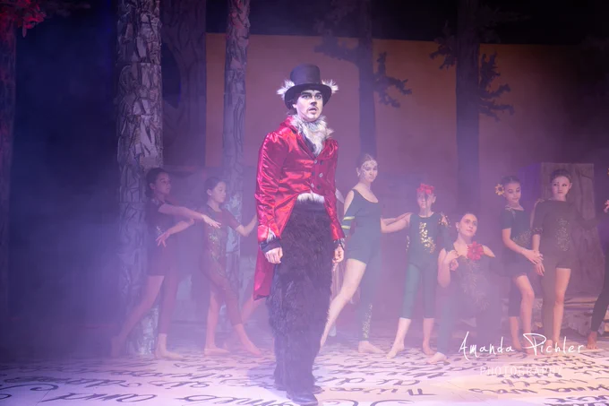 The Big Bad Wolf stands at the front of the stage wearing a red, velvet coat, with smoke and "Into the Woods" dancers behind him.