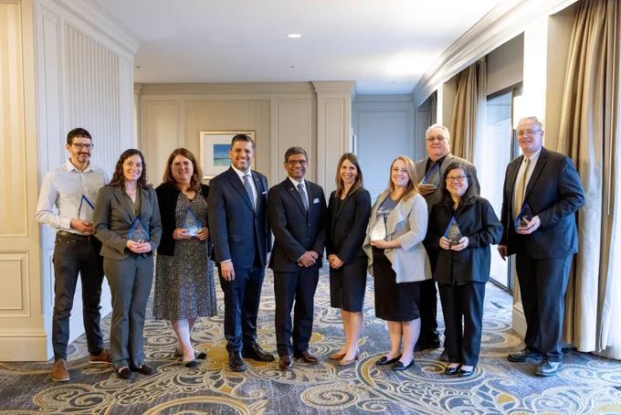 10 people stand together and pose for a photo. Eight of them hold awards.