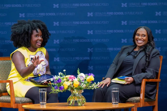Two individuals are seated on stage at the Gerald R. Ford School of Public Policy. They are engaged in conversation, each holding a tablet. A vase of colorful flowers sits on the table between them. The backdrop displays the school's name and logo.