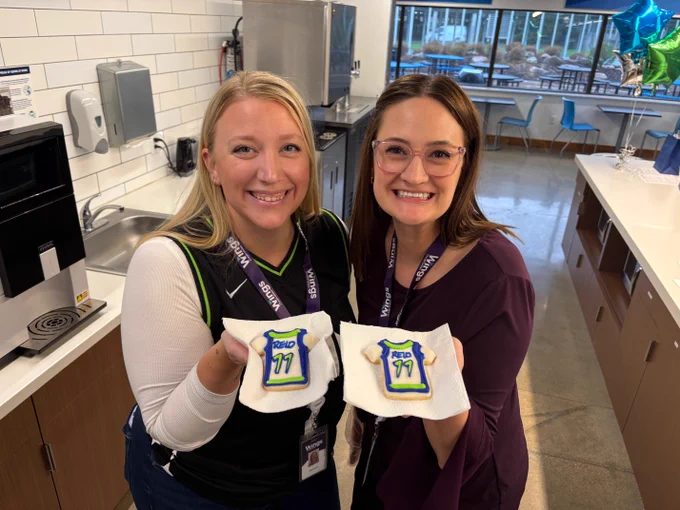 Two people smiling and holding cookies decorated with the word "Voodoo" and the number "77" in a bright kitchen setting.