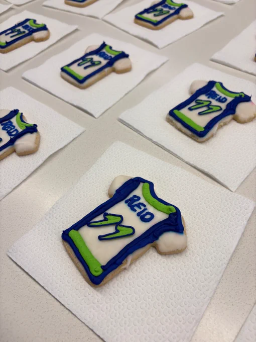 Cookies shaped like sports jerseys are arranged on paper towels. They are decorated with blue, green, and white icing, featuring the name "Reid" and the number "77.