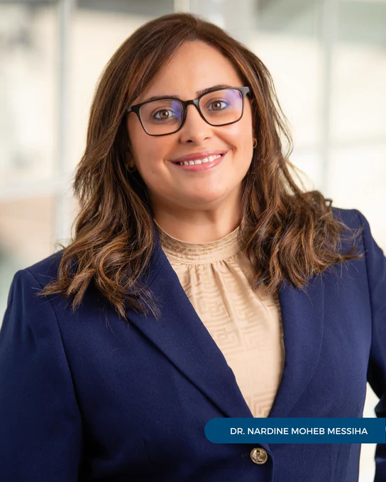 Dr. Nardine Messiha of Tryon Medical Partners smiles and wears a blue blazer and glasses in a professional portrait in front of a paned glass window