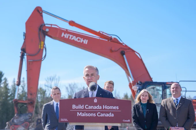 Person speaking a podium with sign “Build Canada Homes | Maisons Canada”, people standing behind. 