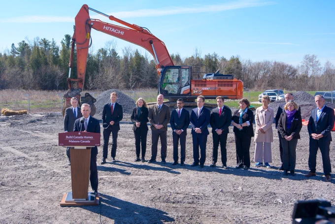 Person speaking a podium with sign “Build Canada Homes | Maisons Canada”, 12 people standing behind. Heavy machinery is positioned in the foreground