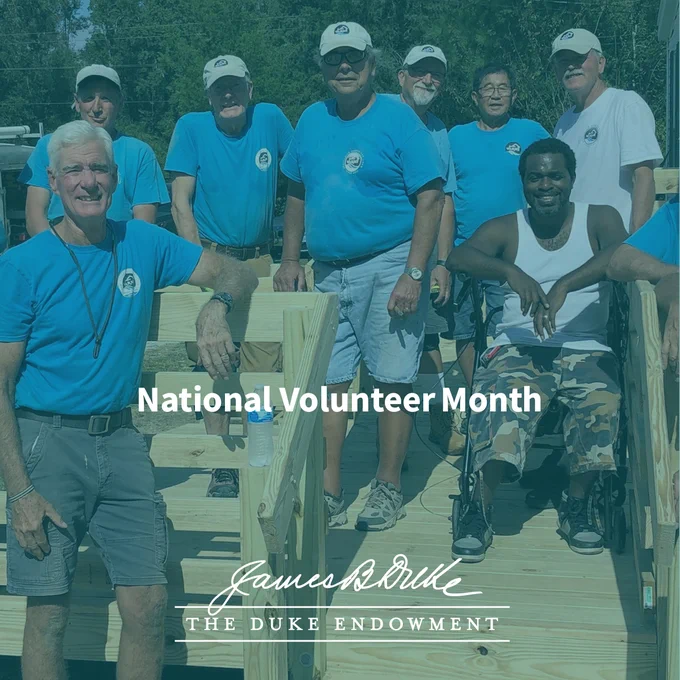 A group of individuals, some standing and one in a wheelchair, wearing matching blue shirts with a logo, pose on a wooden structure outdoors. The image features the text "National Volunteer Month" and "The Duke Endowment" with James B. Duke's signature at the bottom.