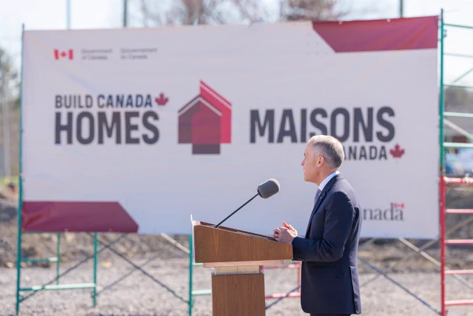 Person speaking at podium with large sign in foreground, “Build Canada Homes | Maisons Canada”