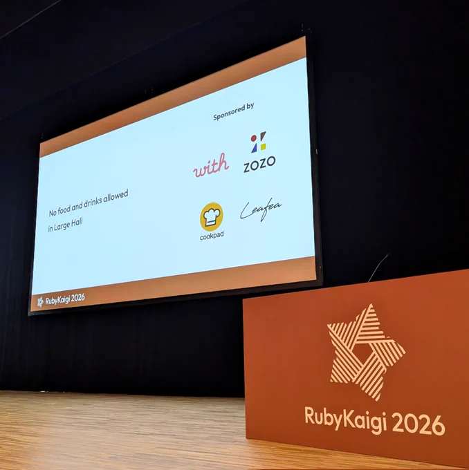 RubyKaigi 2026 メインホール壇上の写真です。スクリーンにスポンサー企業としてクックパッドなどのロゴが表示されています。