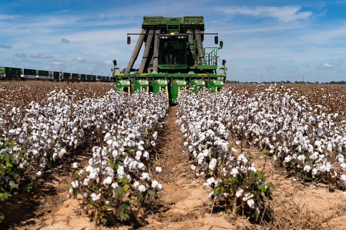 A large green John Deere cotton harvester works in a vast cotton field, with rows stretching into the distance. The sky is clear, and a train is visible on the horizon to the left.