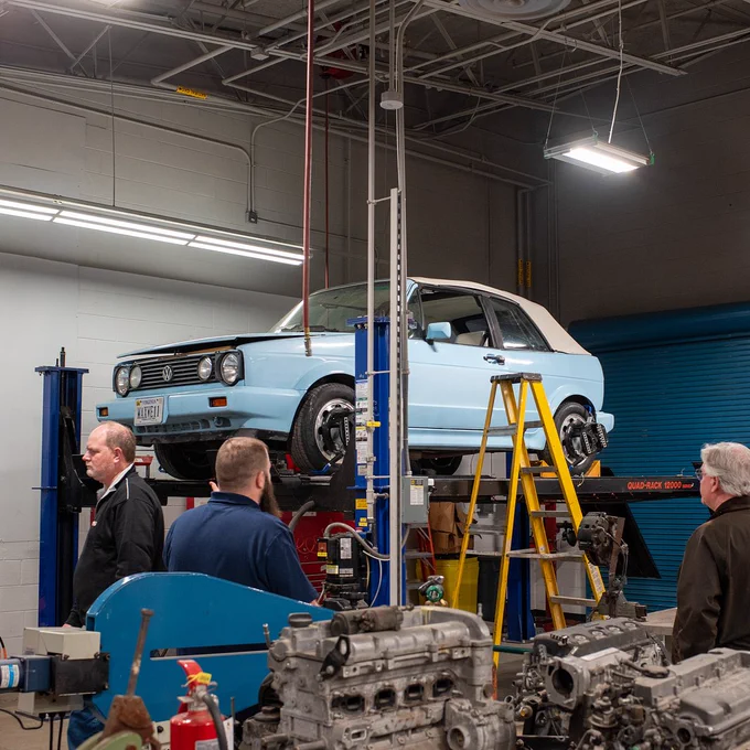 Auto repair shop with a blue hatchback car on a lift, tools and equipment scattered on the floor, and several people inspecting the car.
