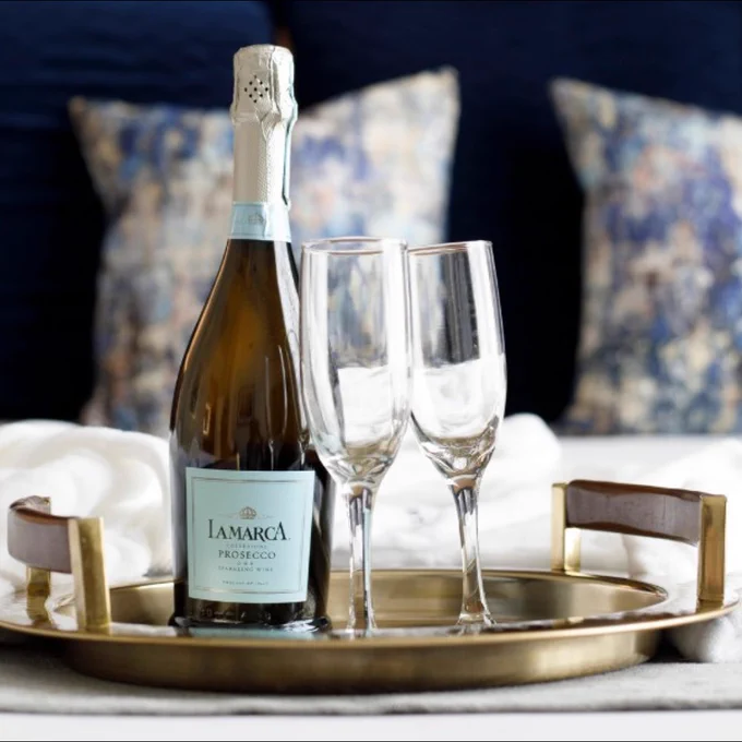 A bottle of La Marca Prosecco is placed on a round tray alongside two empty champagne flutes. The tray sits on a bed with patterned pillows in the background.