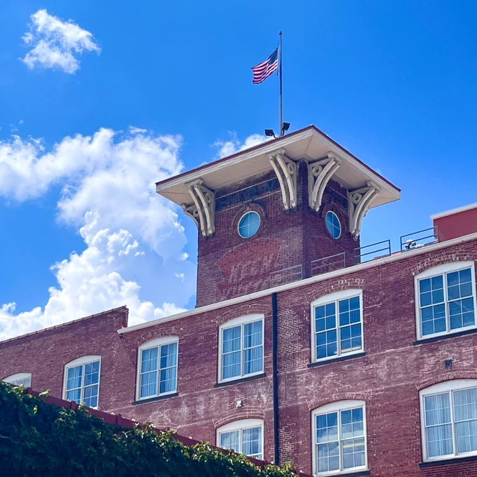 A brick building with a prominent tower under a bright blue sky with fluffy clouds. An American flag is atop the tower. The building displays faded painted signs with "Whitney Lofts" visible.