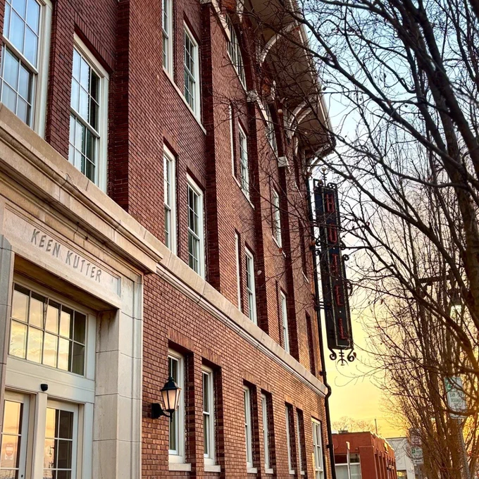 A brick building with "Keen Kutter" etched above an entrance, featuring a vertical "HOTEL" sign. The scene includes bare trees and a sunset sky.