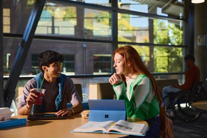 Two people sitting at a table in a bright, modern space, one holding a drink and the other using a Microsoft Surface device.