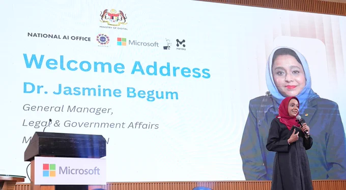 Speaker delivering welcome address at Microsoft event with presentation slide behind.