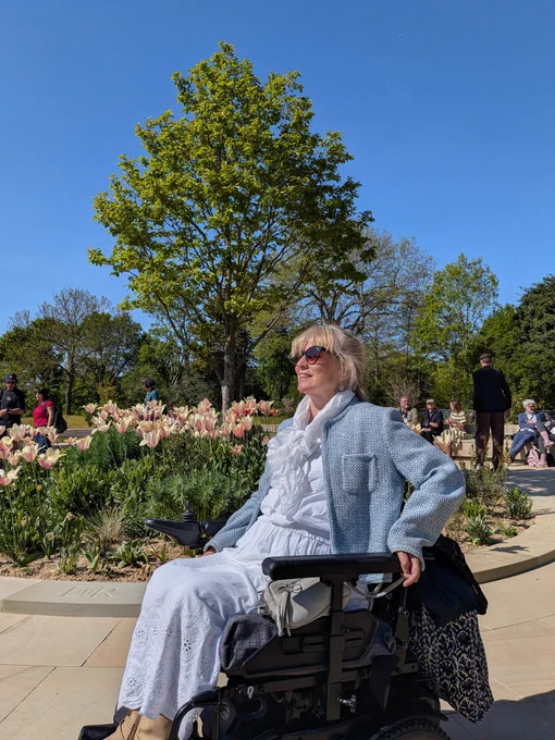 Clare using a power chair on a wide white path in QE2 gardens. Tulips flower in the bed behind here