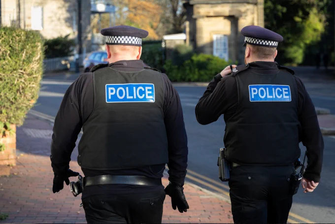 Two police officers in uniform walking away from the camera on a street.