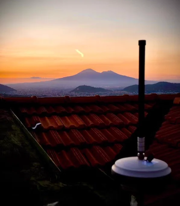 Wingbits receiver antenna on a rooftop overlooking Mount Vesuvius at sunset, symbolizing community-built infrastructure connecting a local commons to a global aviation data network.