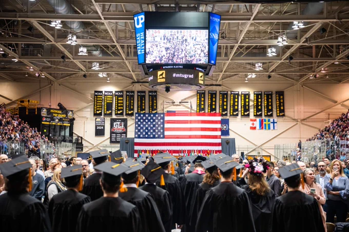 Michigan Tech graduates in caps and gowns face a large American flag hanging in a packed indoor arena during a commencement ceremony.