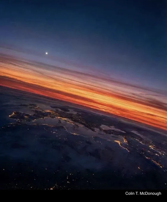 A breathtaking high-altitude view of the Earth at twilight. A vibrant orange and red sunset glows along the horizon, fading into a deep indigo sky where a single, bright planet—Venus—shines clearly. Below the clouds, the lights of coastal towns and cities outline the land near the English Channel. The photographer’s credit, Colin T. McDonough, is visible in the bottom right corner.