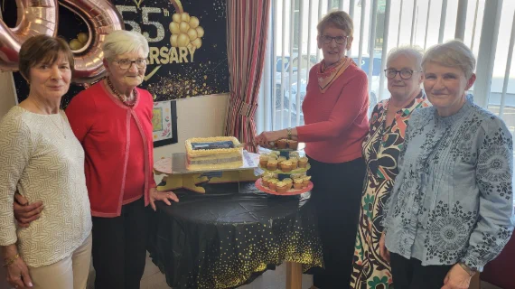 Five women at the anniversary celebration, standing around a table with a cake and cupcakes.