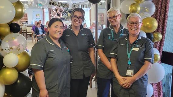 Four smiling care assistants in grey uniform pictured at the anniversary celebration with balloons and a banner in the background.