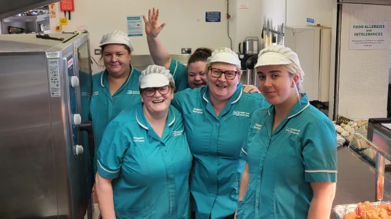 A group of six smiling catering staff in teal uniforms and white caps pose for a photo in a kitchen.
