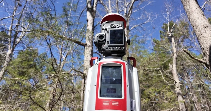 A robotic scanner stands center of the image surrounded by trees with a blue sky above.