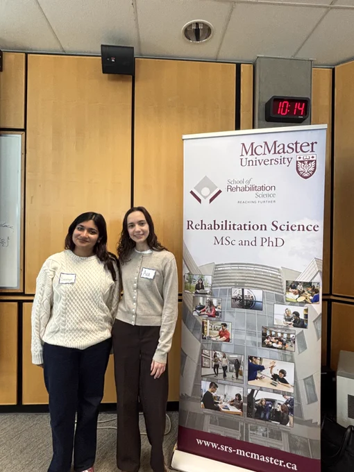AHA! members Saanya (left) and Ava (right) standing next to the McMaster Rehab Science banner